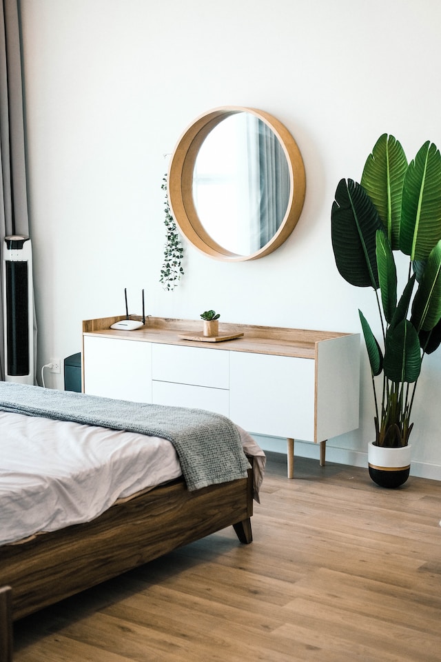 A modern bedroom with a wooden bed frame, white and wood sideboard, round mirror, and potted plant. The room appears clean and well-maintained, showcasing key furnishings and conditions relevant for a rental inspection.