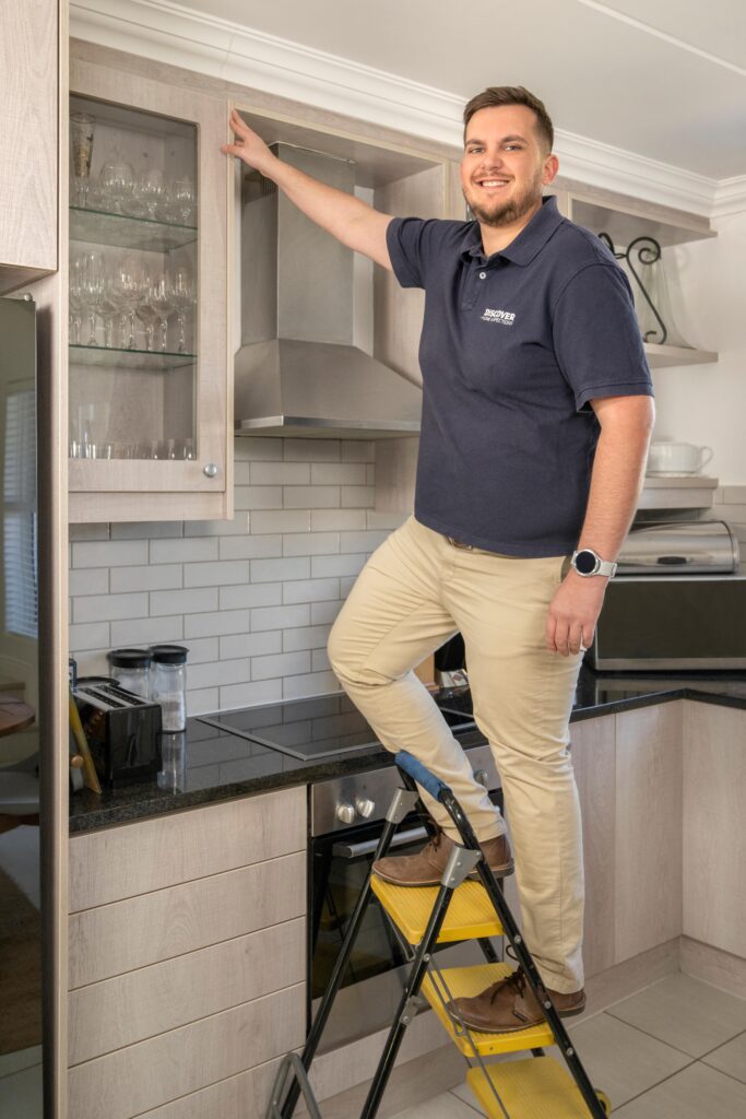 A man standing on a ladder pointing toward a possible defect in a modern kitchen. He is wearing a navy blue short-sleeved polo shirt with the logo "Discover Home Inspections" on the chest.