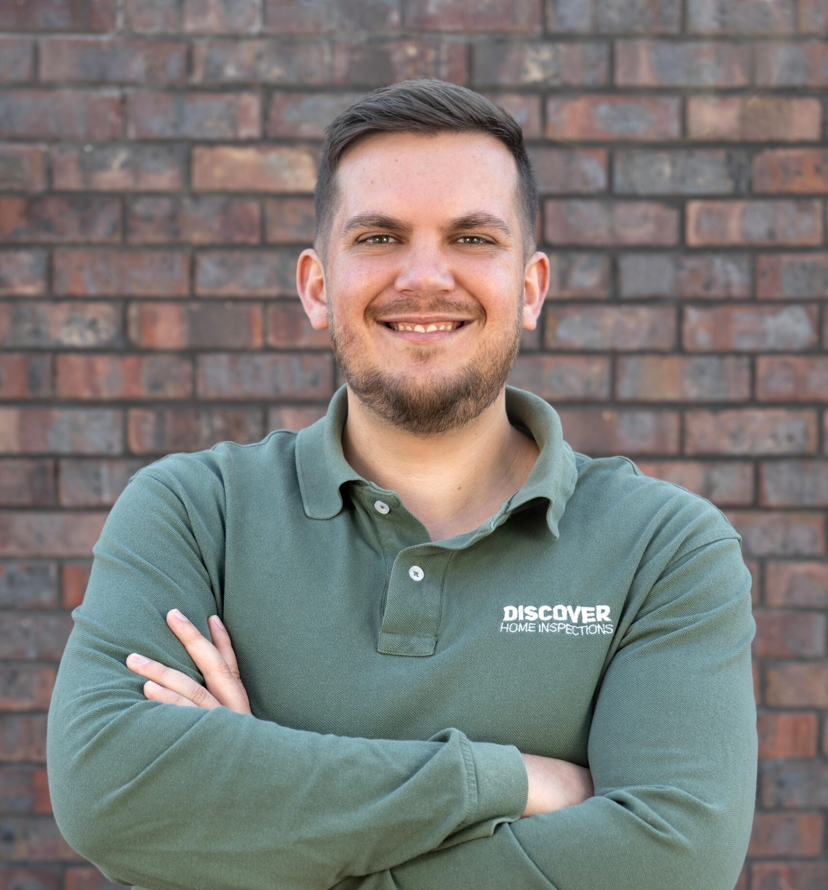 A man with short hair and a beard is smiling and standing in front of a brick wall. He is wearing a green long-sleeved polo shirt with the logo "Discover Home Inspections" on the chest. His arms are crossed, and he appears confident and approachable.